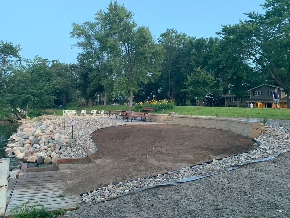 Lakeside landscape with gravel area, picnic table, and trees at dusk.