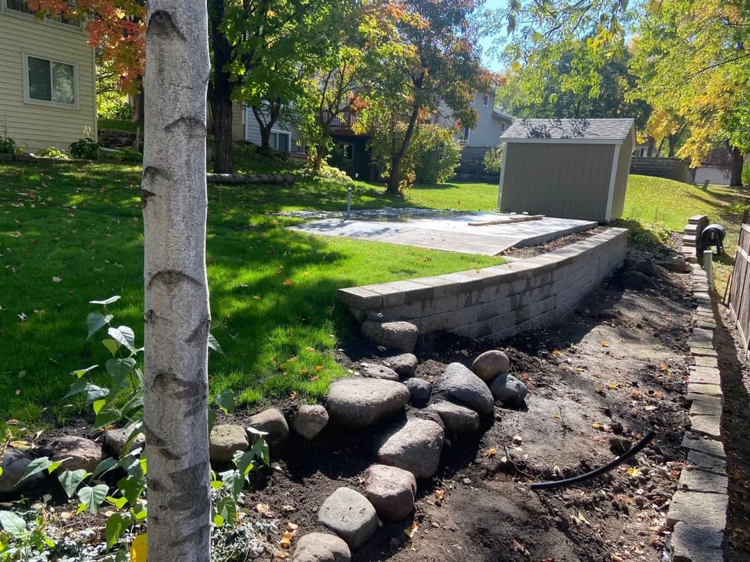 Landscaped backyard with stone wall, green lawn, and shed surrounded by autumn trees.