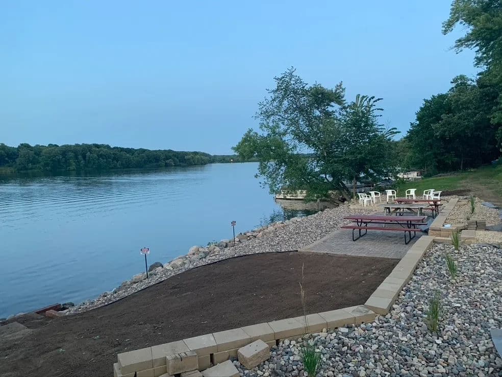 Peaceful lakeside scene with a stone patio, picnic tables, and seating by the water's edge.