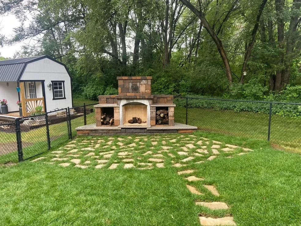 Backyard outdoor fireplace surrounded by a stone patio and green grass in a serene setting.