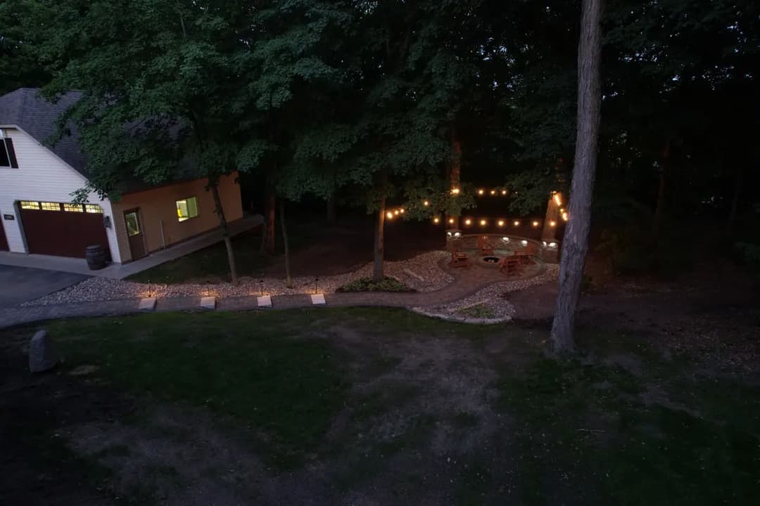 Cozy outdoor living space with string lights, fire pit, and stone pathway surrounded by trees.
