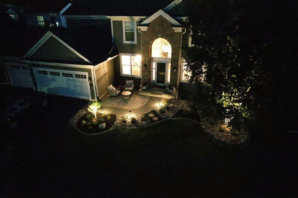 Aerial view of a well-lit house at night with a stone patio and illuminated garden.