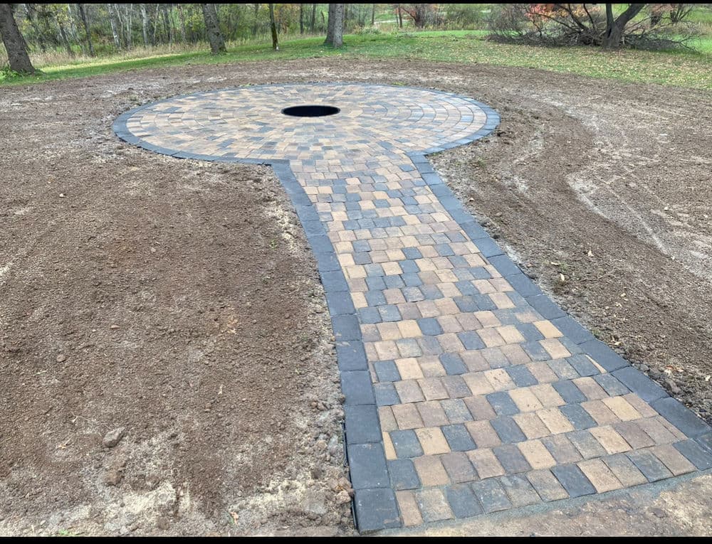 Circular stone patio with a central hole, surrounded by a dirt area and trees in the background.