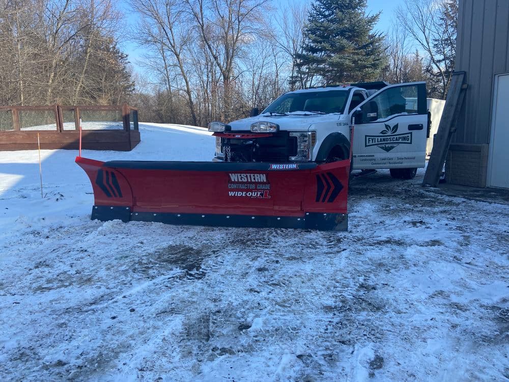 Western snow plow attached to a white truck in a snowy landscape with pine trees.