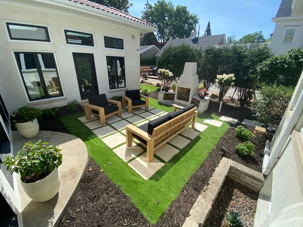 Stylish patio with modern seating, green turf, and a cozy fireplace surrounded by lush landscaping.