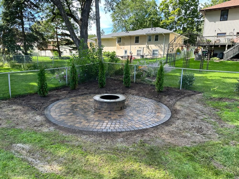 Patio area with circular stone fire pit surrounded by landscaping in backyard setting.