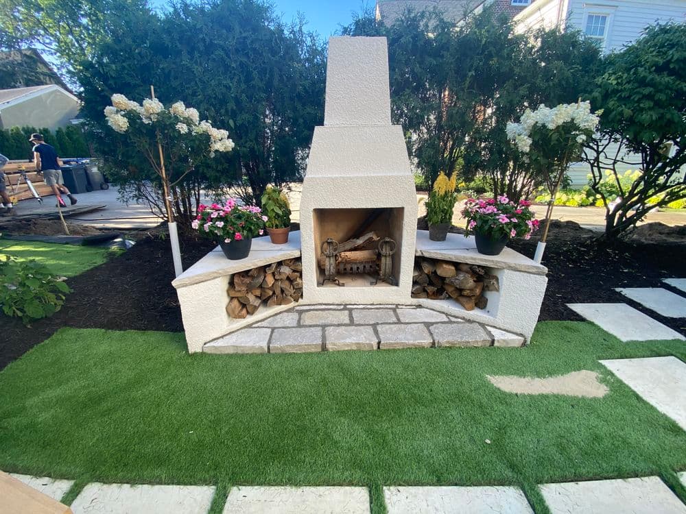 Outdoor fireplace with stacked wood, surrounded by flower pots and landscaped garden.