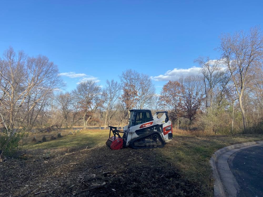 Bobcat skid steer near cleared land with trees under a blue sky and scattered clouds.